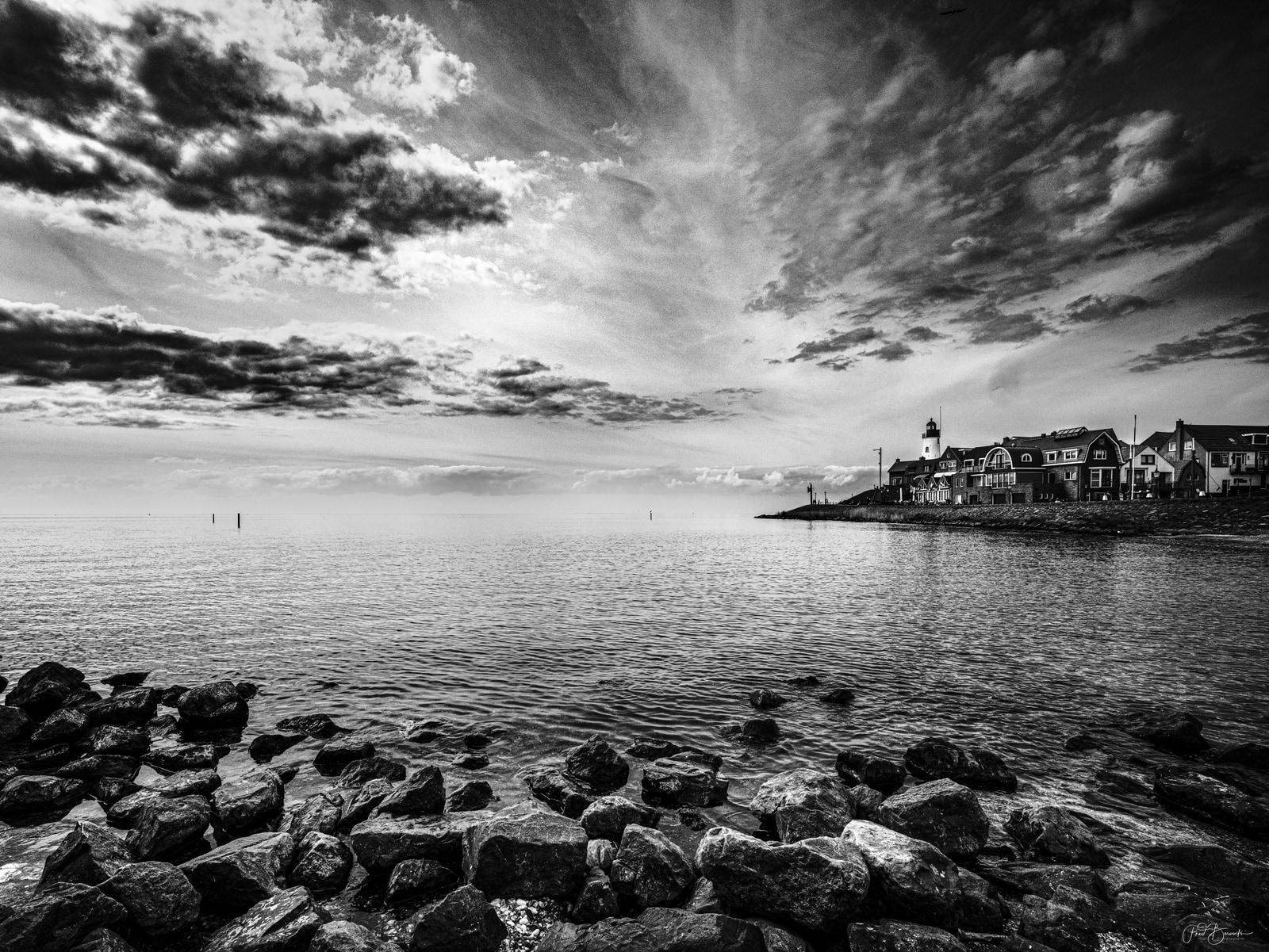 A rocky shoreline faces a calm body of water under a dramatic, brooding sky, with a distant building perched on a small island.