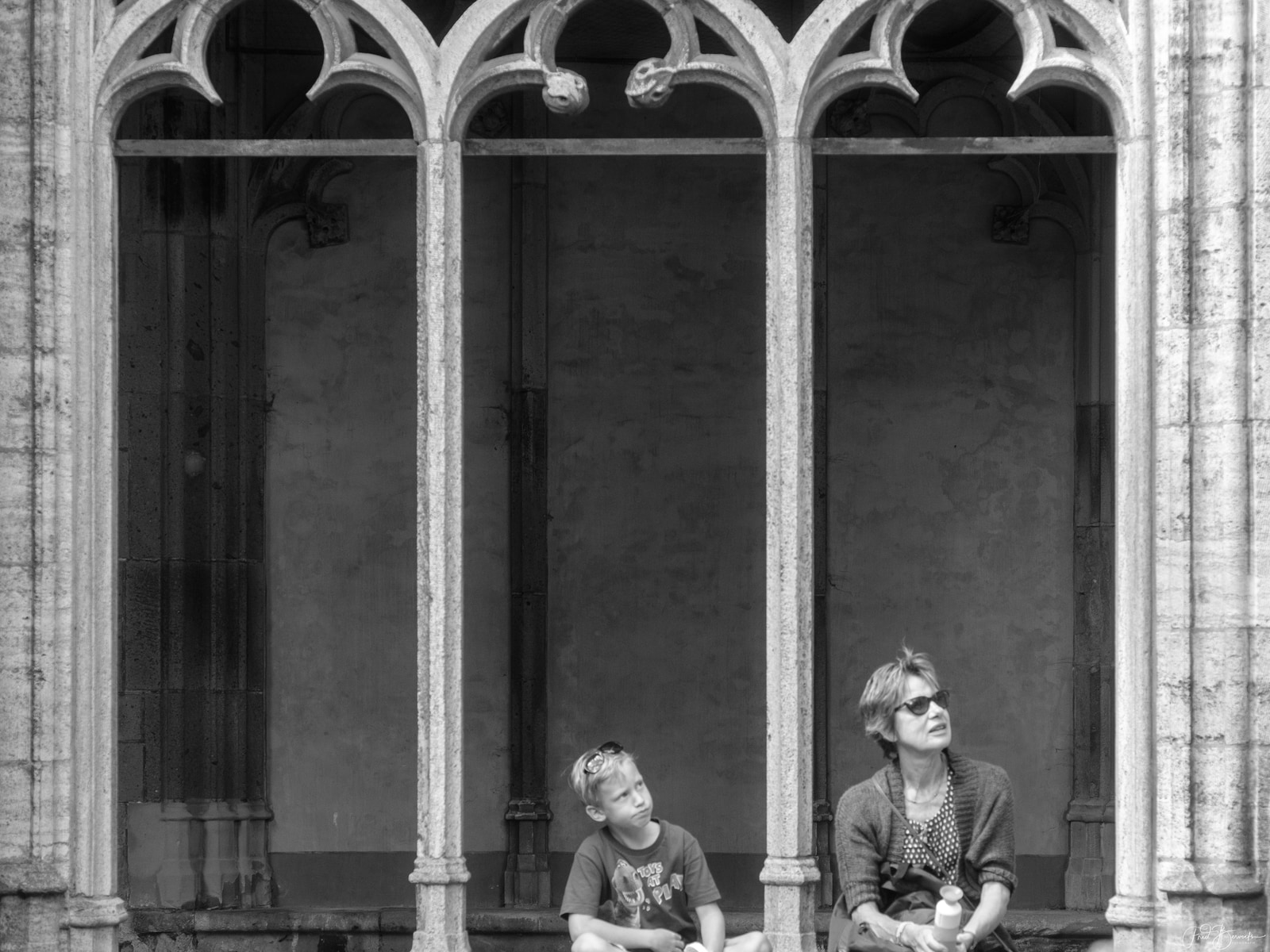 Two people sit side by side on a stone ledge in front of an ornate arched window.