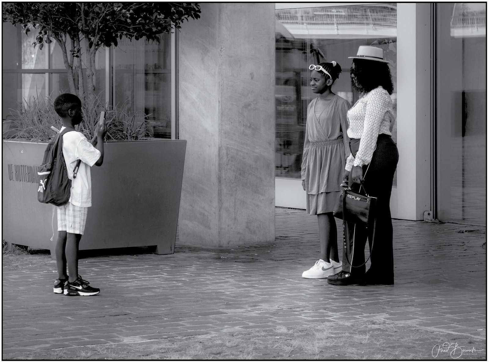 Three people stand together on a paved surface near a building entrance, with one person holding a camera.
