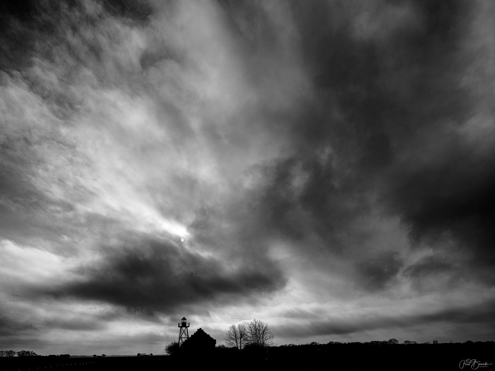 A dramatic black-and-white landscape shows a solitary water tower rising against a brooding, cloud-filled sky.