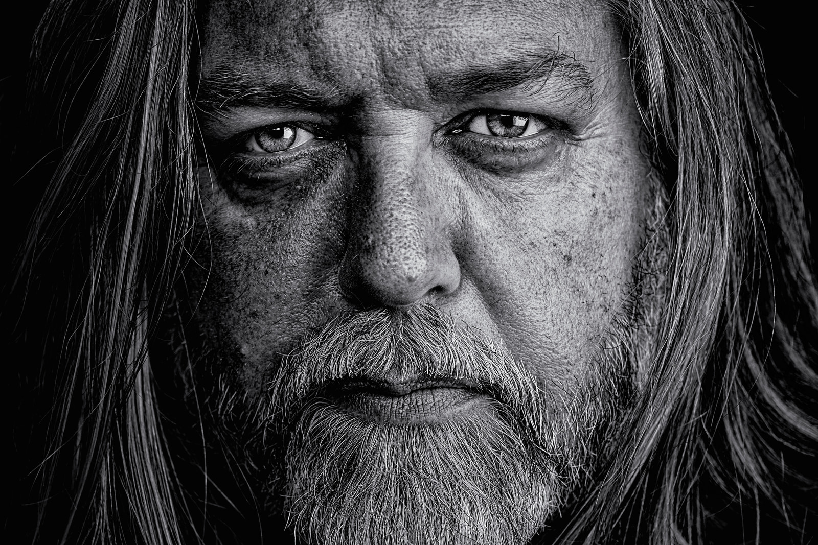 A close-up portrait of a man with long hair and a beard, captured in black and white.