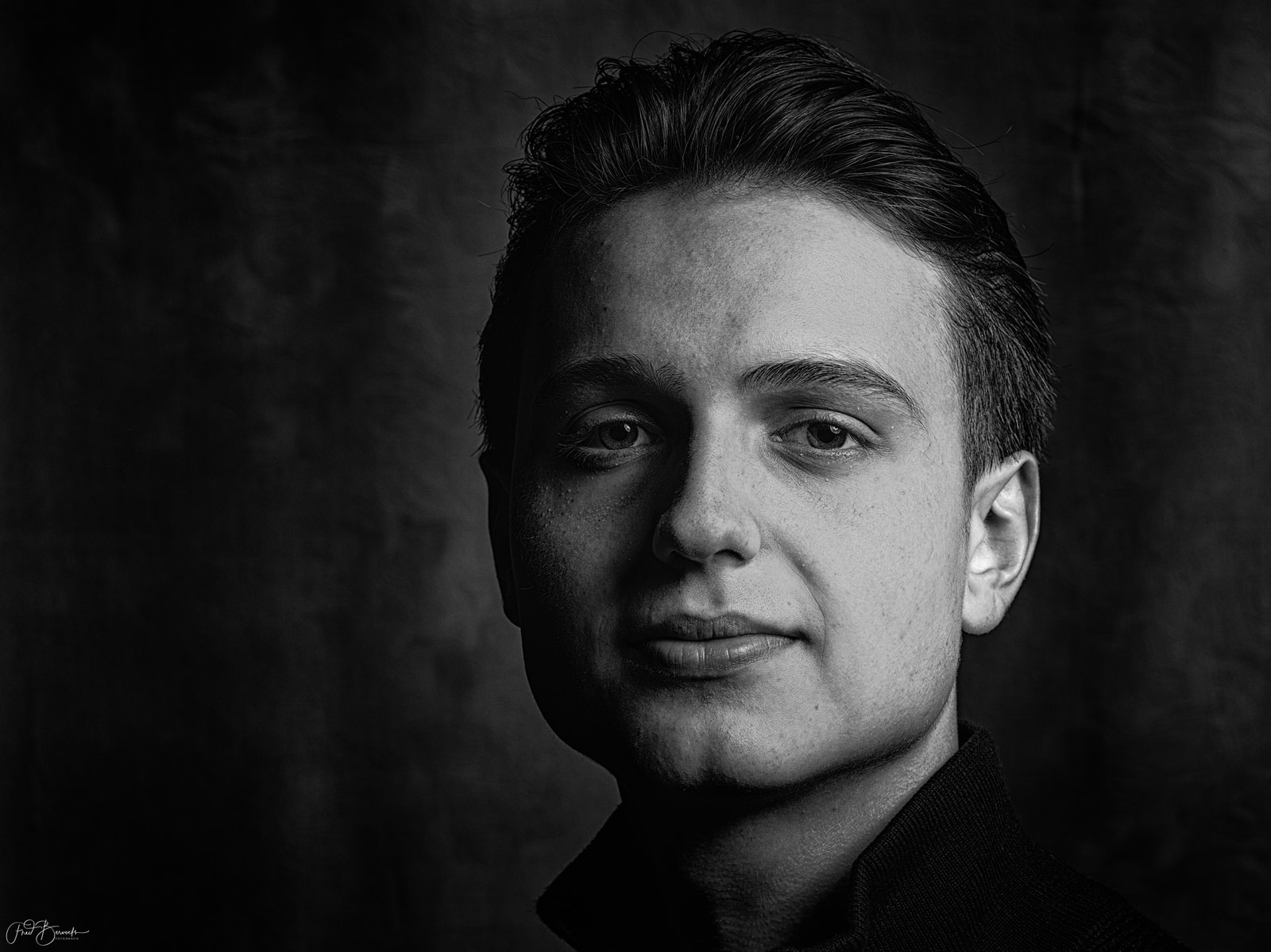 A man with short hair and a slight smile is shown in a close-up portrait against a dark, textured background.