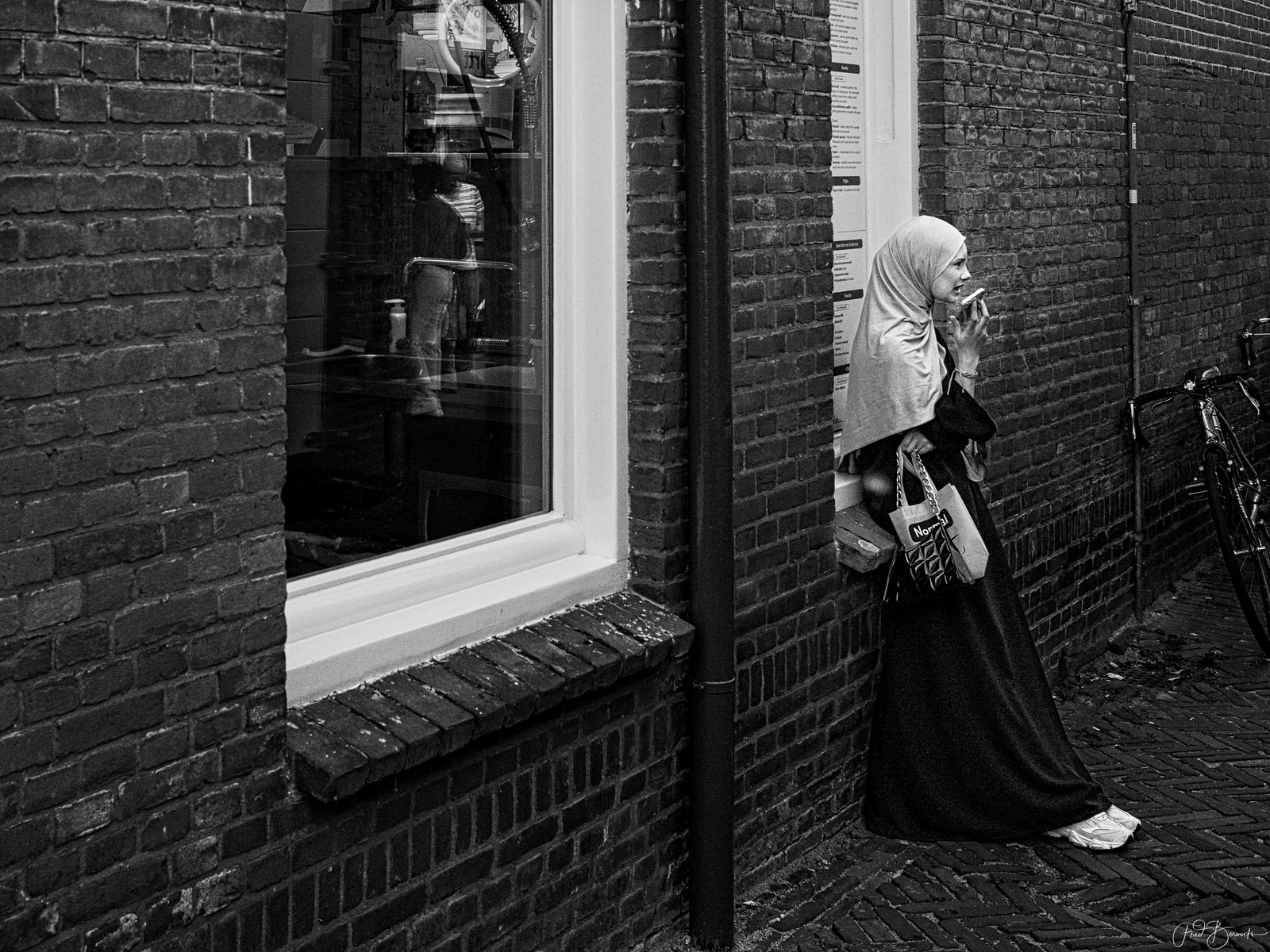 A woman in traditional attire stands near a brick building, smoking a cigarette while holding a bag.