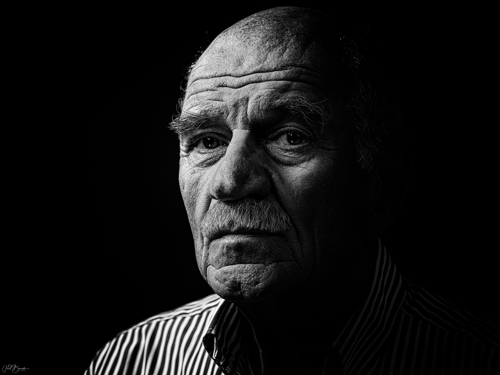 An older man with a mustache and short hair is shown in a close-up portrait against a dark background.