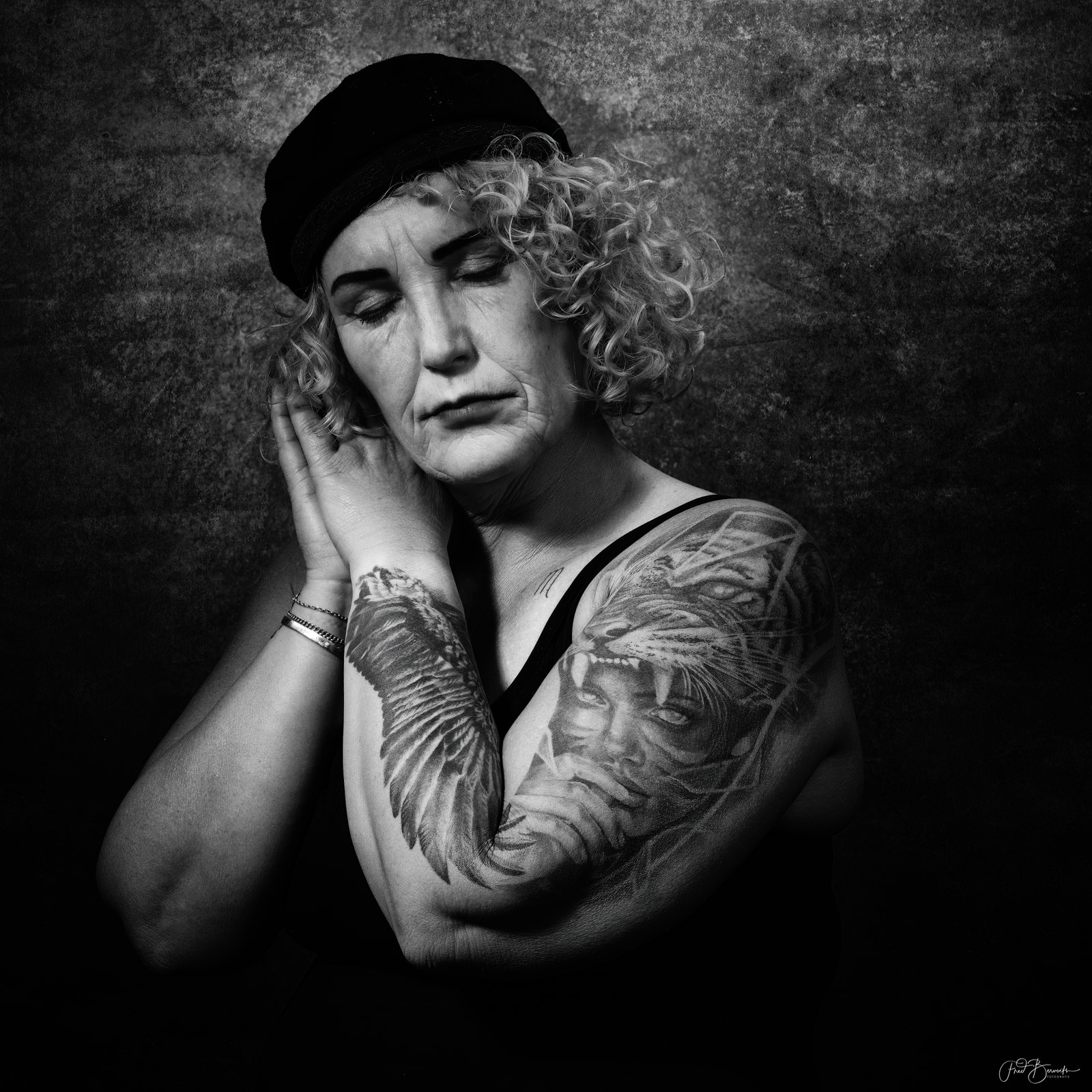 A person with curly hair and a beanie sits with one hand on their cheek, wearing a sleeveless top that reveals large, detailed tattoos on both arms.