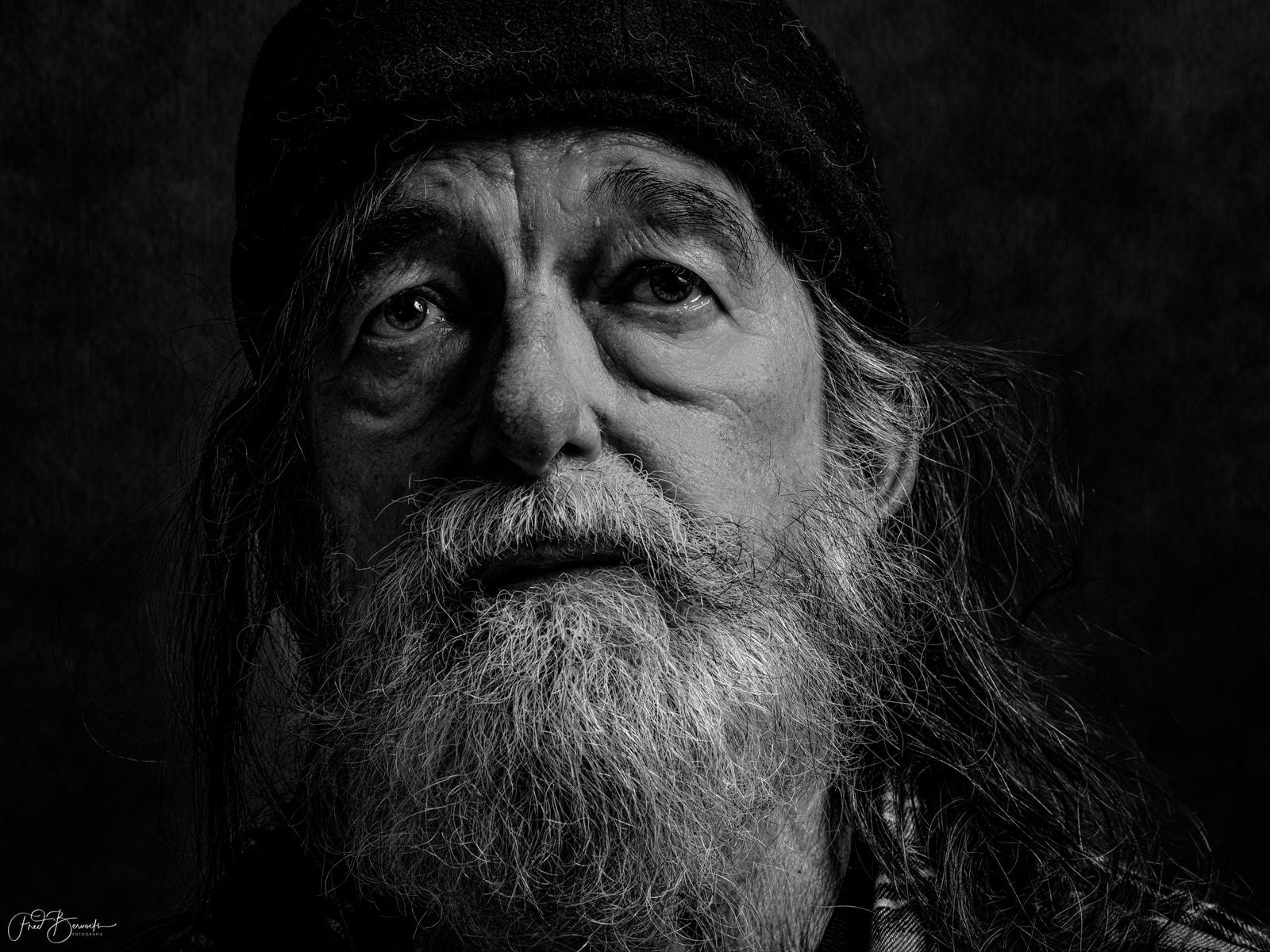 A man with long hair and a beard gazes directly at the camera against a dark background.