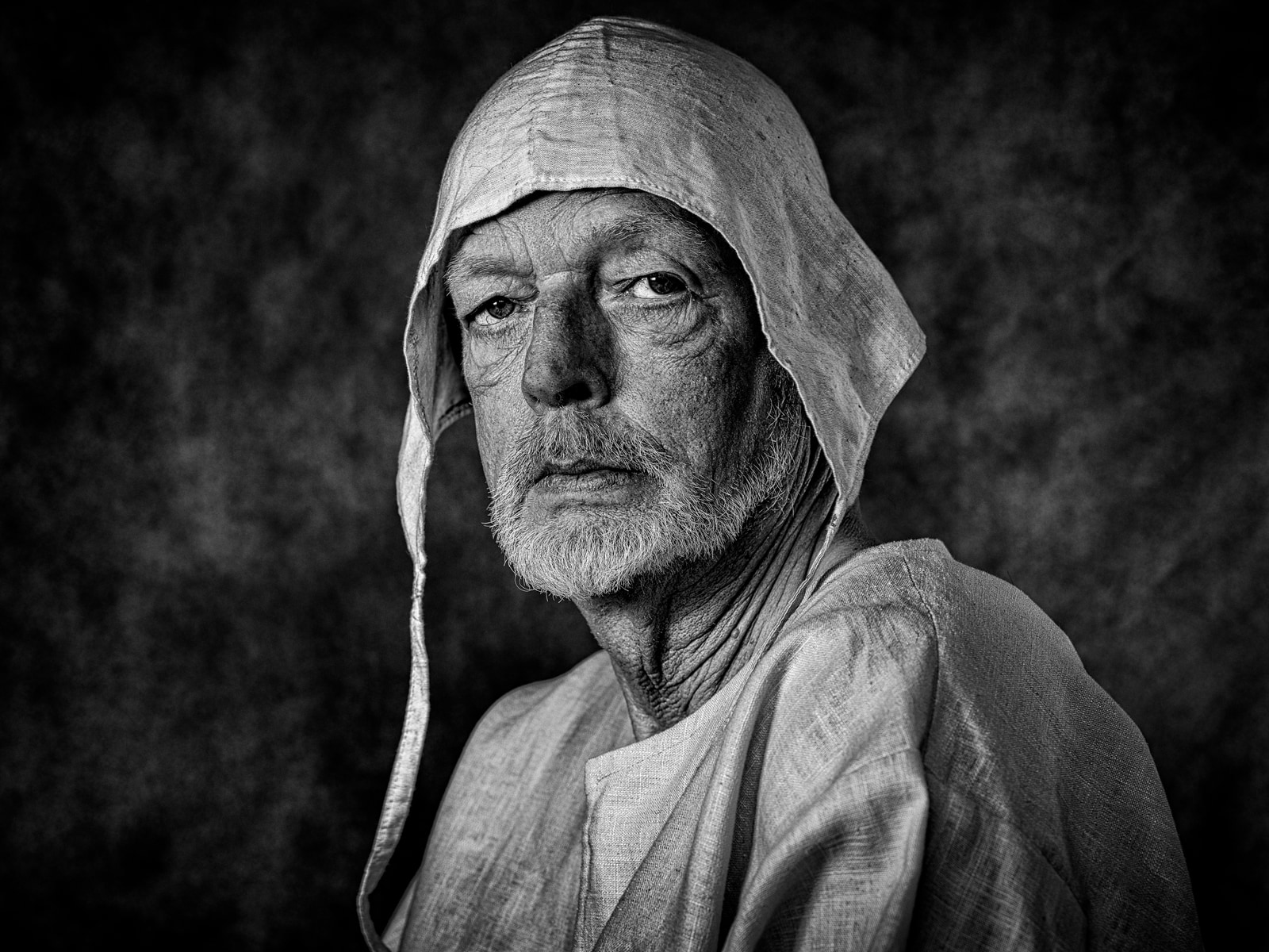 An elderly man with a beard and a hooded garment gazes directly at the camera.
