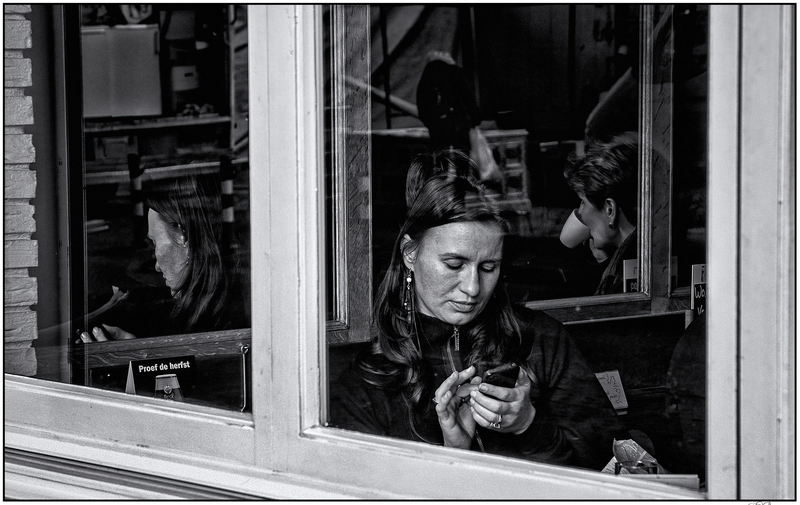 A woman sits inside a café, looking down at her phone, with another person visible through the window behind her.