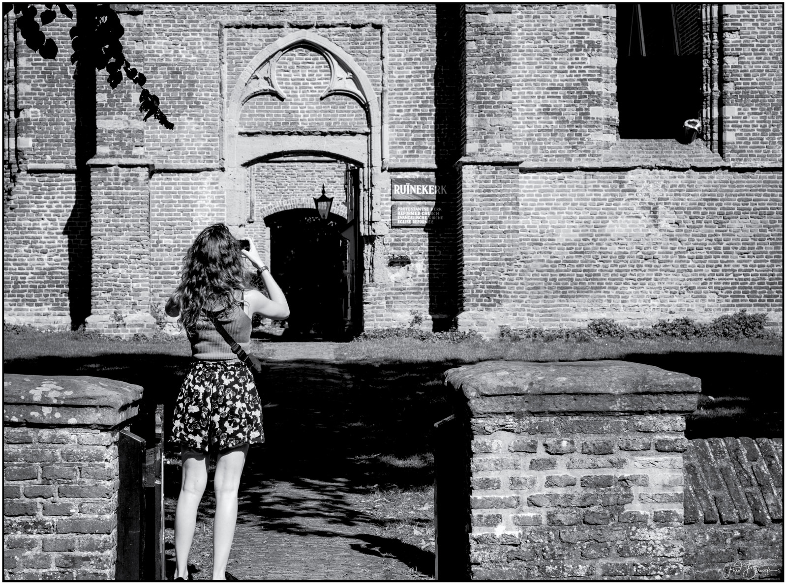 A woman stands near a brick building entrance, facing away from the camera and looking at her phone.