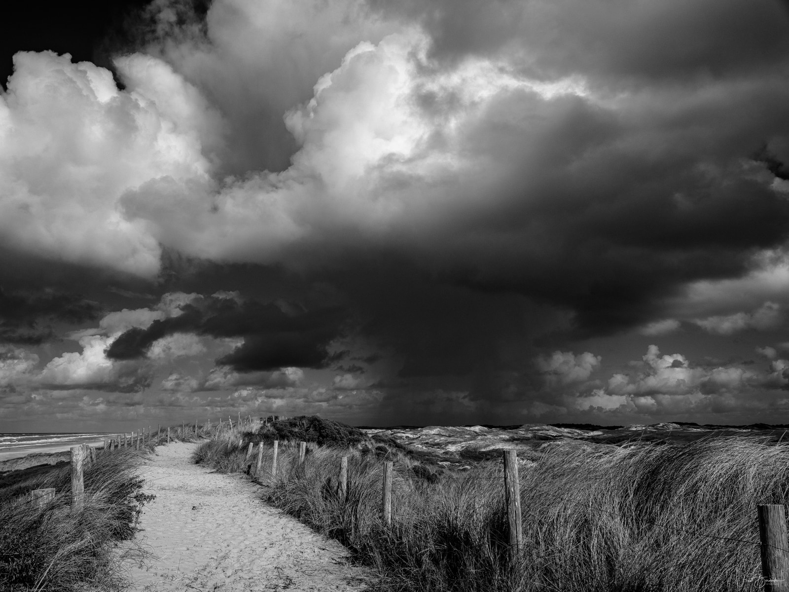 A sandy beach path winds through tall grasses under a dramatic, stormy sky with dark, billowing clouds.