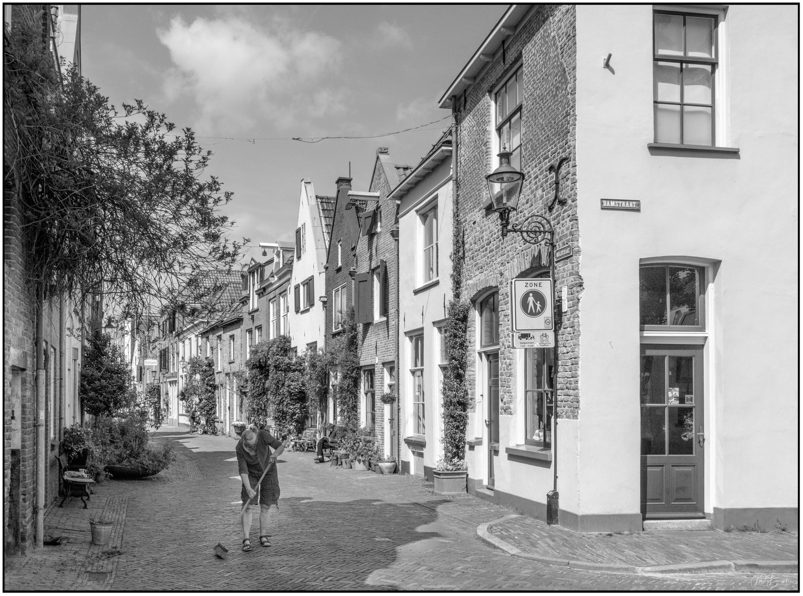 A person sweeps a cobblestone street lined with traditional brick houses and street lamps.