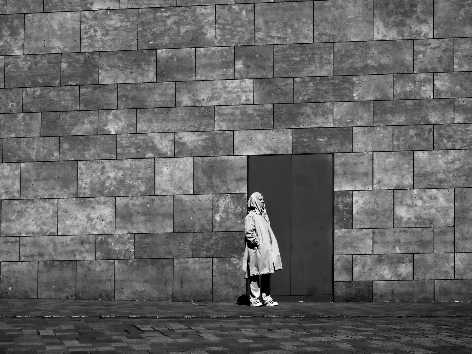 A solitary figure stands in a doorway against a large, textured concrete wall.