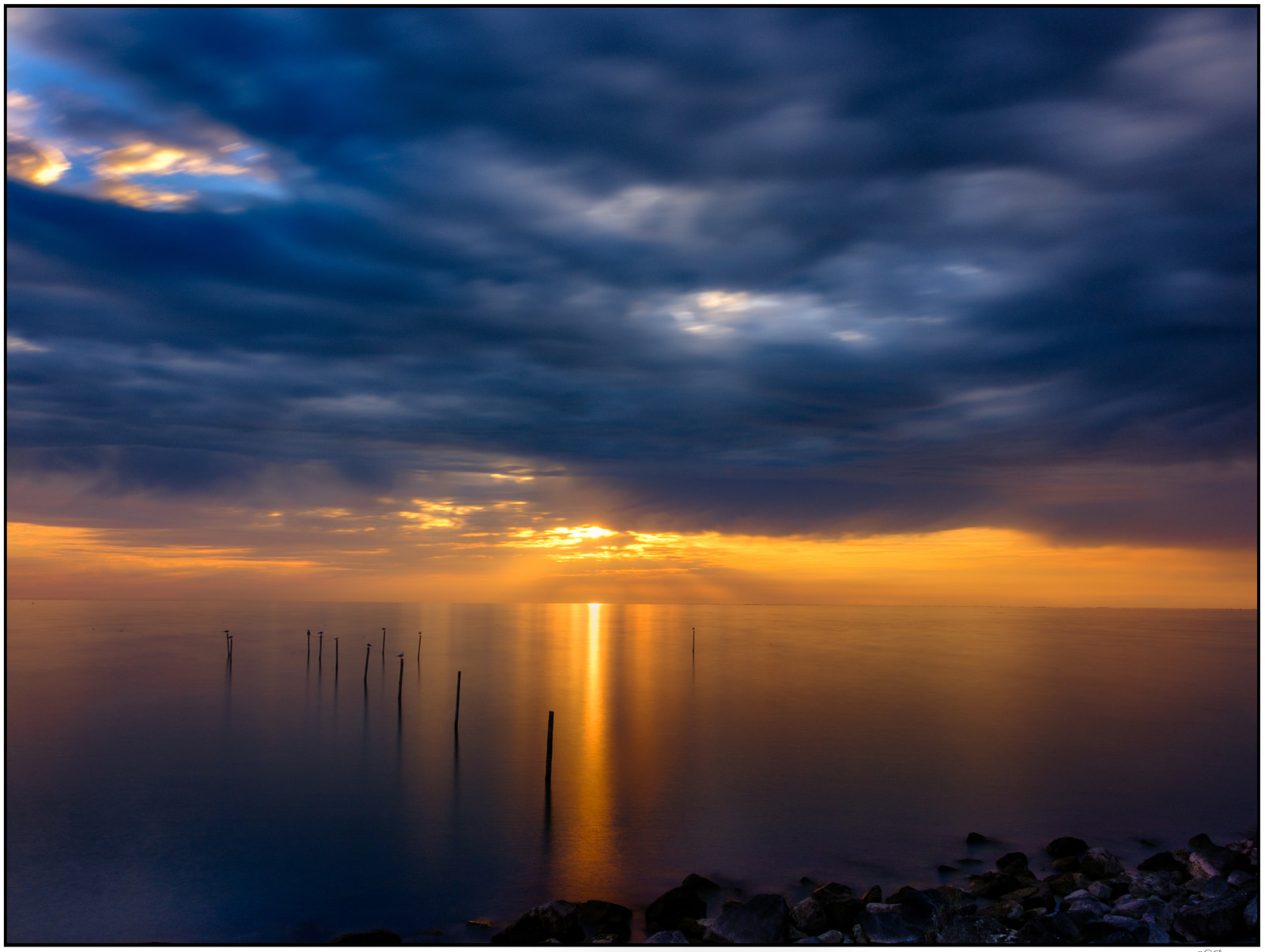 A tranquil seascape at sunset with the sun low on the horizon, casting a warm golden reflection across calm water, and a line of wooden posts standing upright in the shallow foreground.