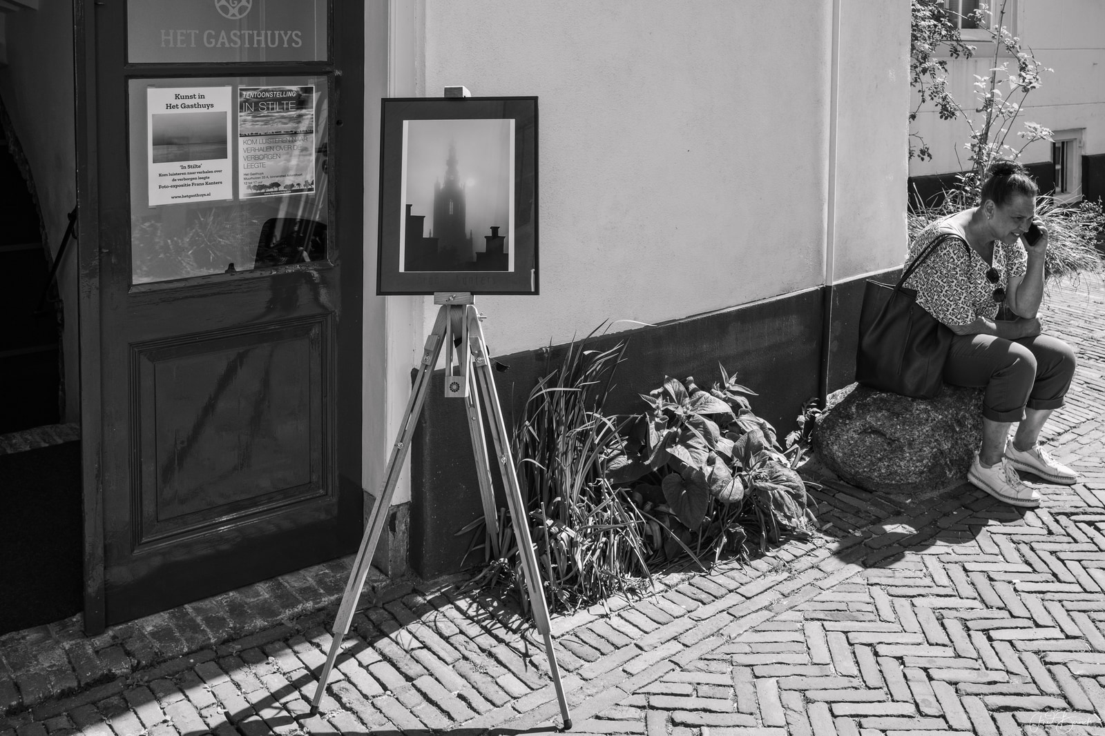 A woman sits on a stone bench near a doorway, with a framed artwork on a tripod beside her.