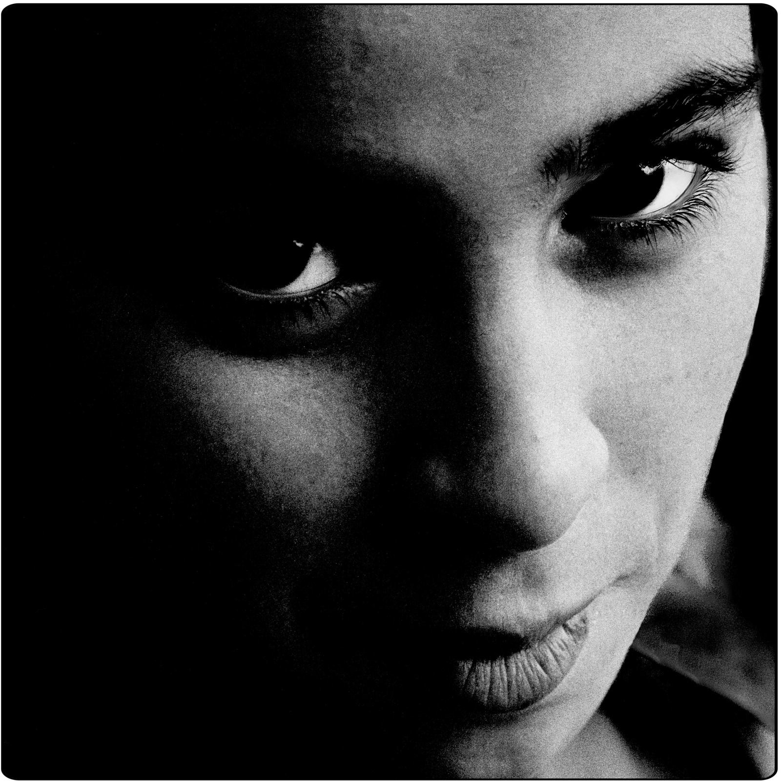 A close-up view of an eye and part of the surrounding face, captured in black and white with strong contrast and deep shadows.
