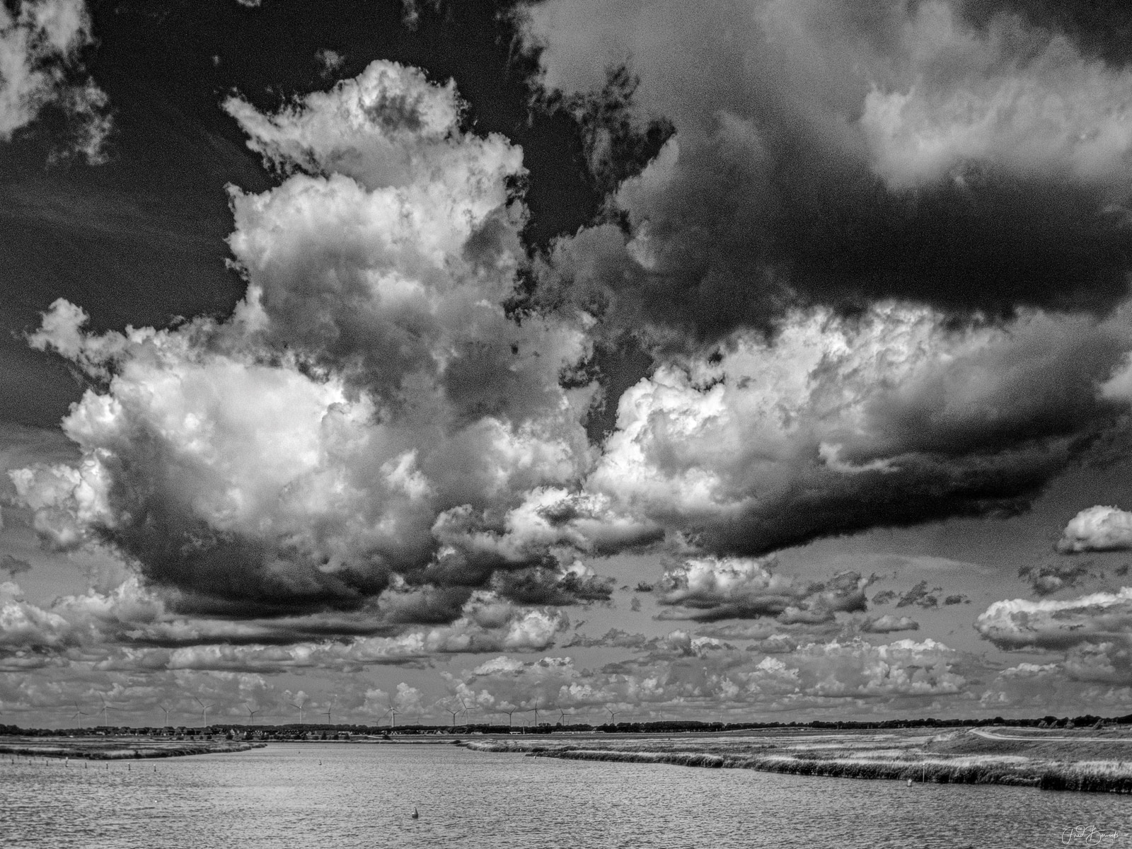 A dramatic black-and-white landscape shows towering, billowing storm clouds over a calm body of water, with distant wind turbines visible on the horizon.
