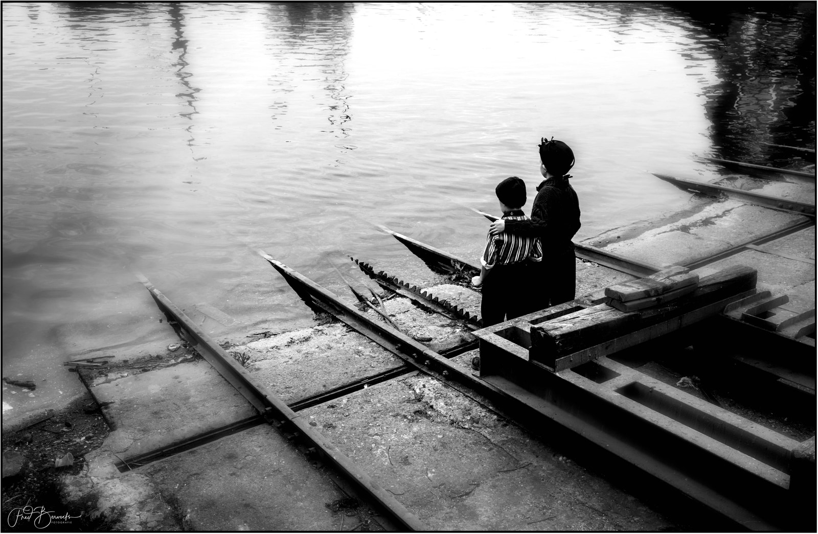 Two children stand together on a weathered dock, gazing out over calm water.