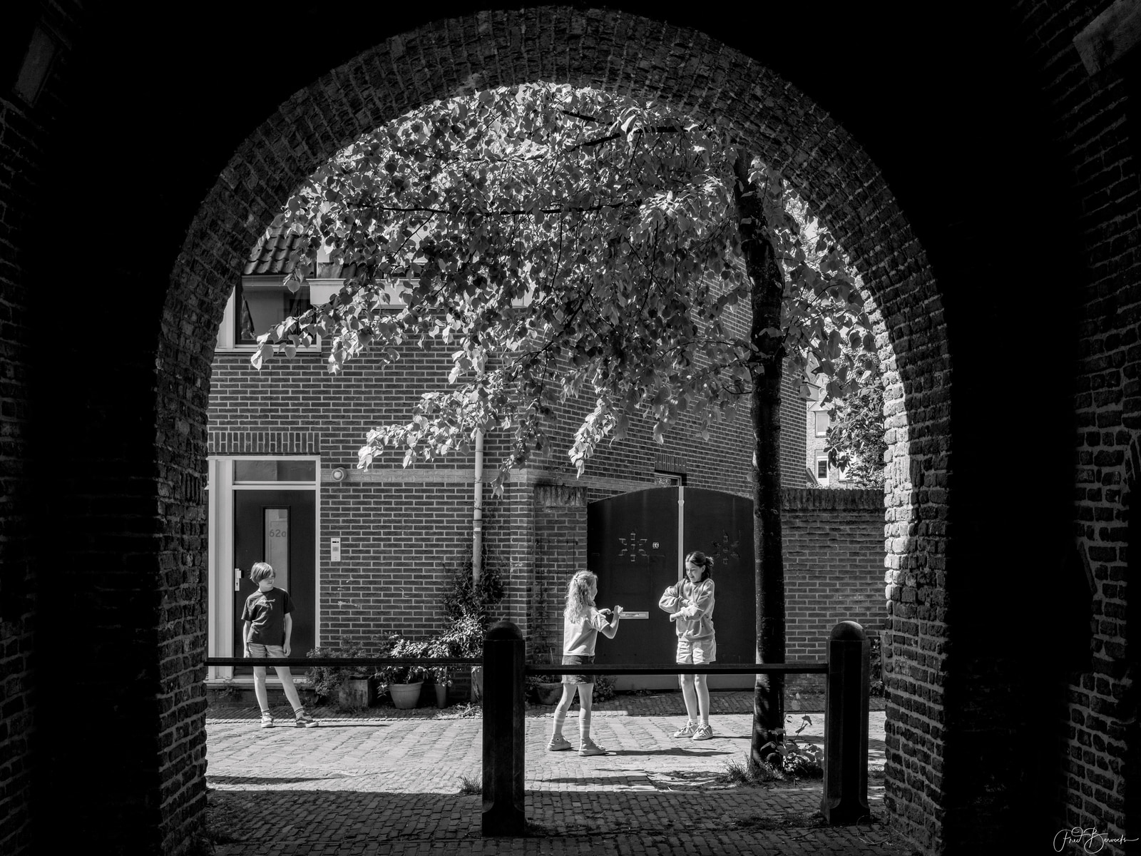 Three children stand near a brick archway, with one leaning against the wall and two others facing each other, framed by a tree's canopy.