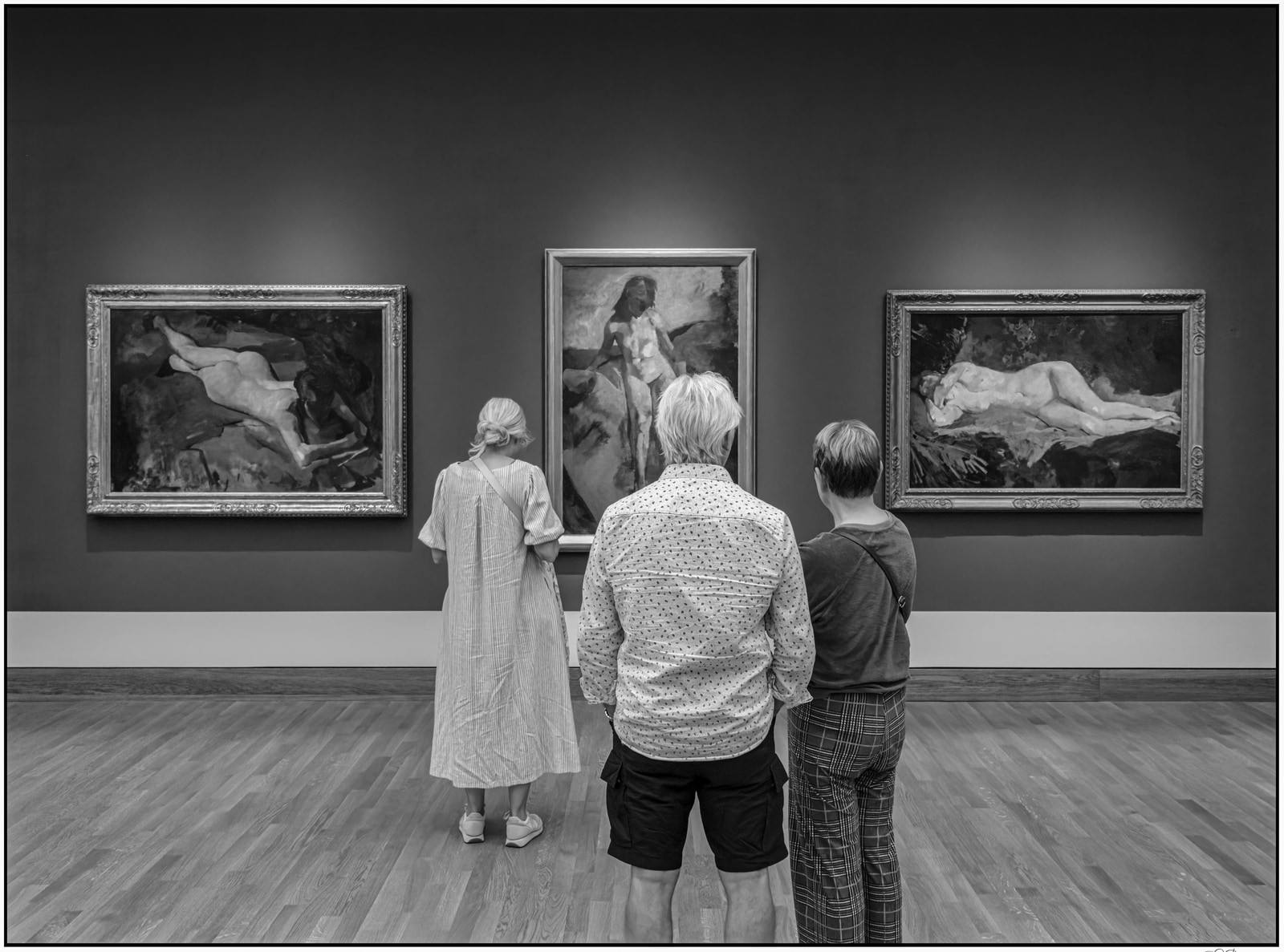 Three people stand together observing framed artworks in a gallery space.