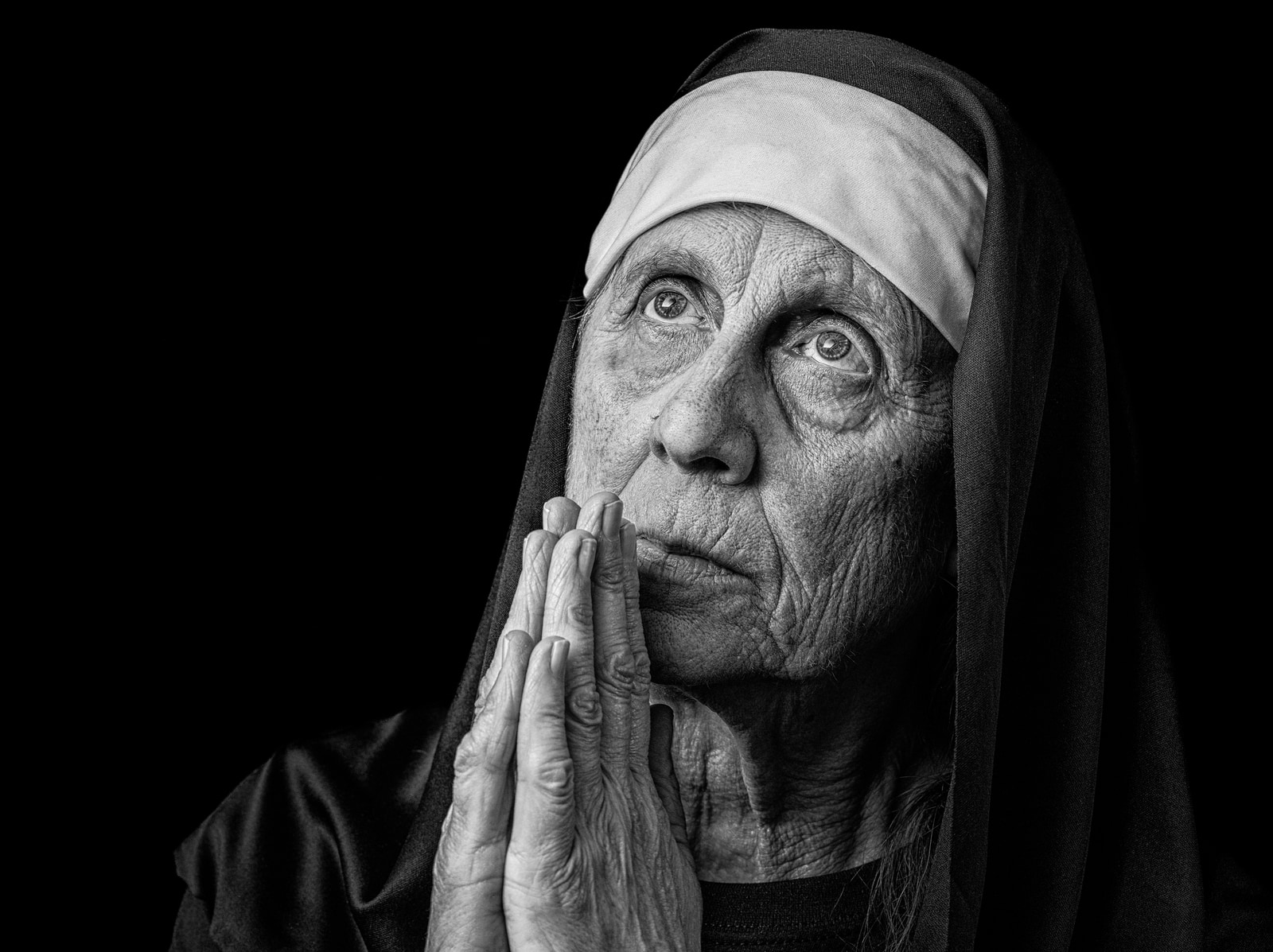 An elderly person with a head covering has their hands together in a prayer position against a dark background.