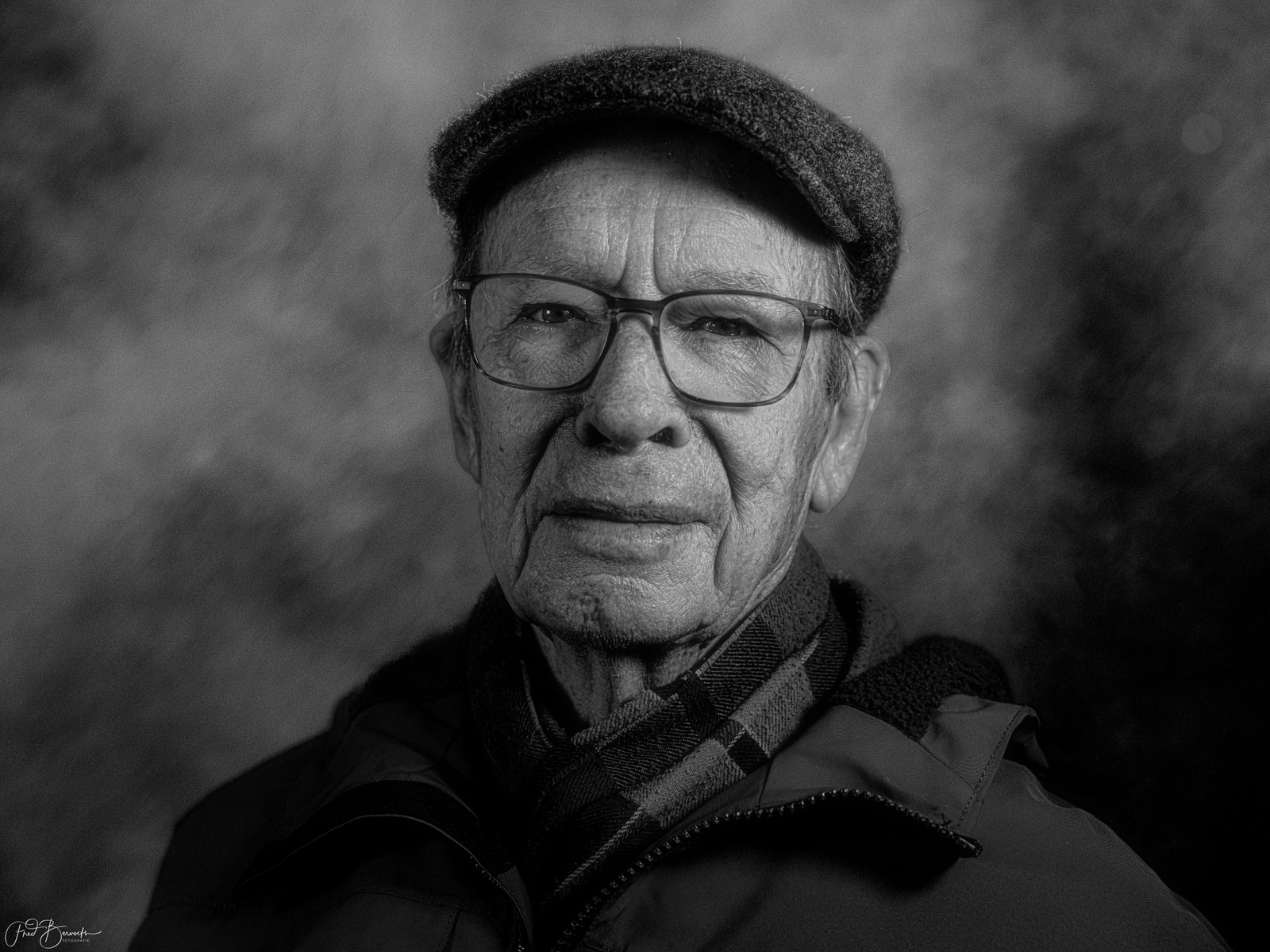 An older man with glasses and a hat looks directly at the camera.