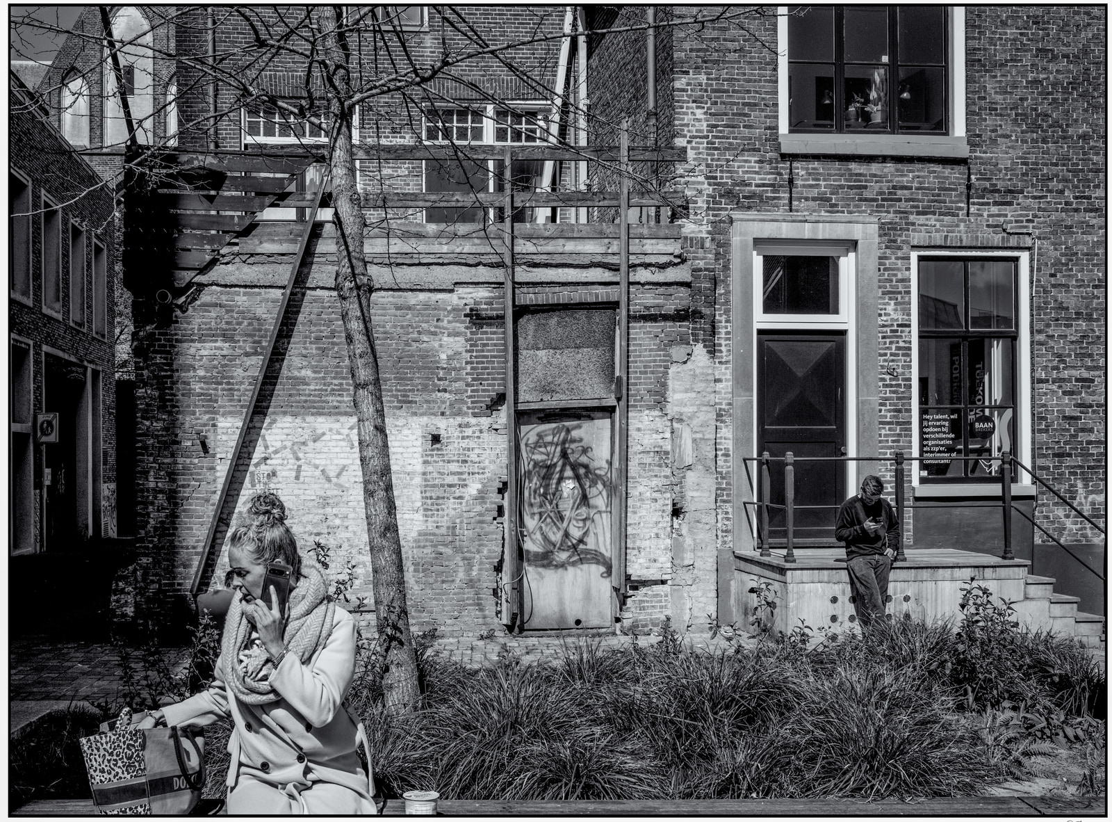 A woman in a coat uses a phone while walking past a weathered brick building with graffiti on a closed door and a small garden in front.