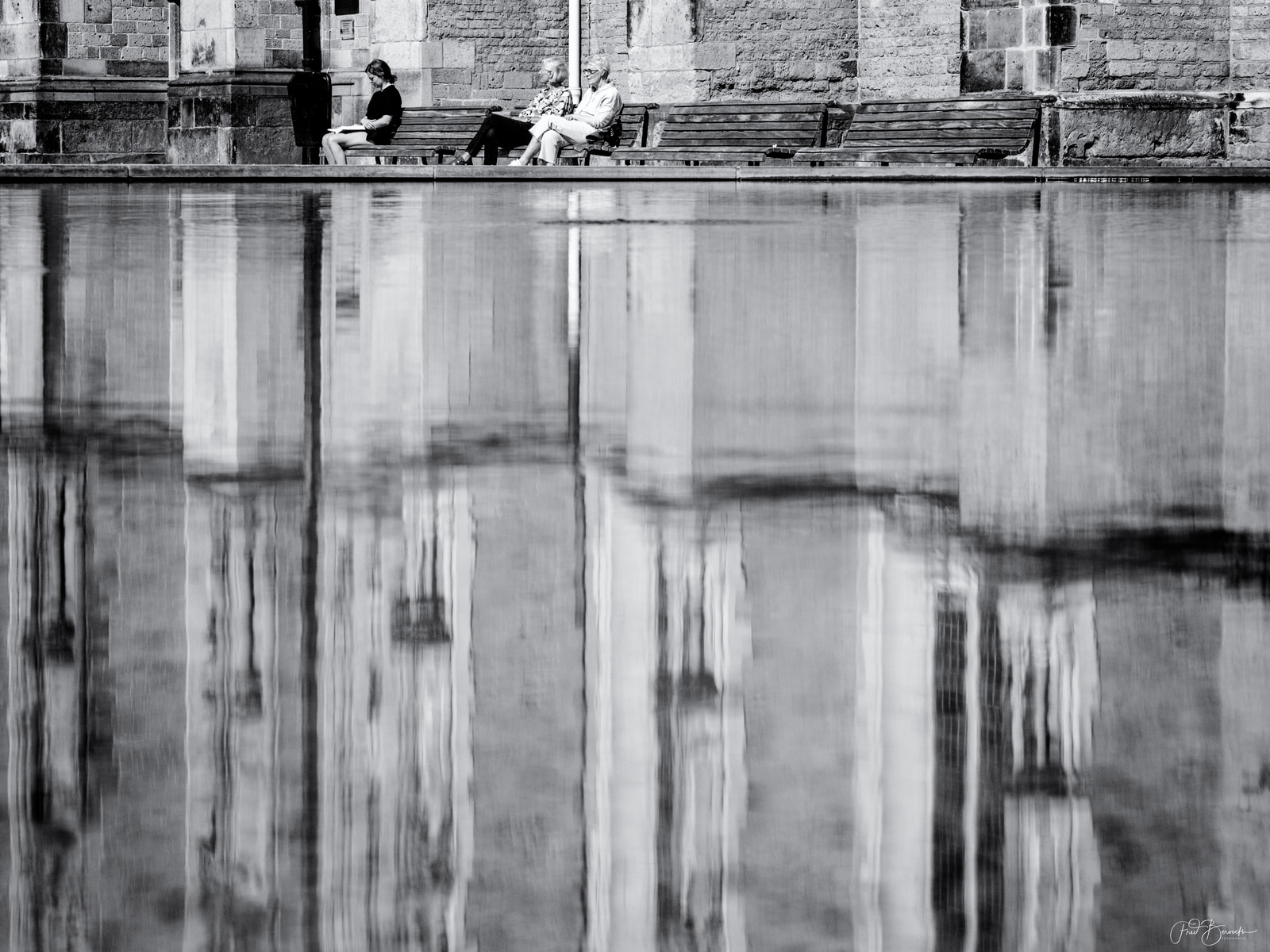 Two people sit on a bench beside a reflective pool, with a stone building reflected in the water.