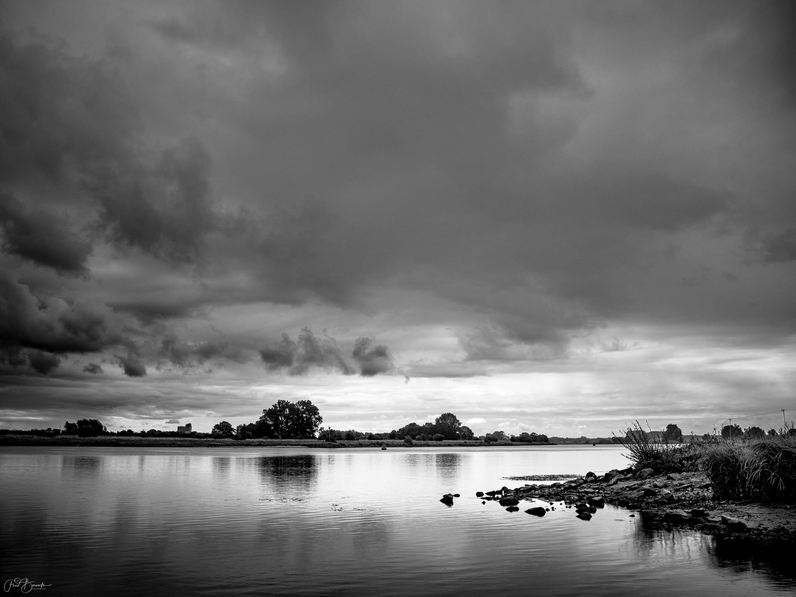 A wide, calm river stretches under a dramatic, brooding sky filled with thick, dark clouds; the far bank is lined with trees and distant structures.