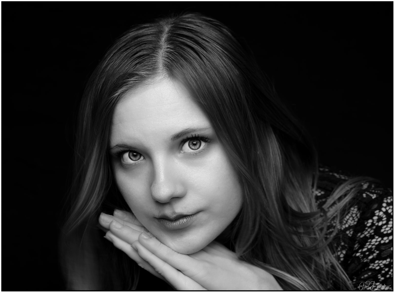A young woman with long hair gazes directly at the camera, her hand gently touching her cheek against a dark background.