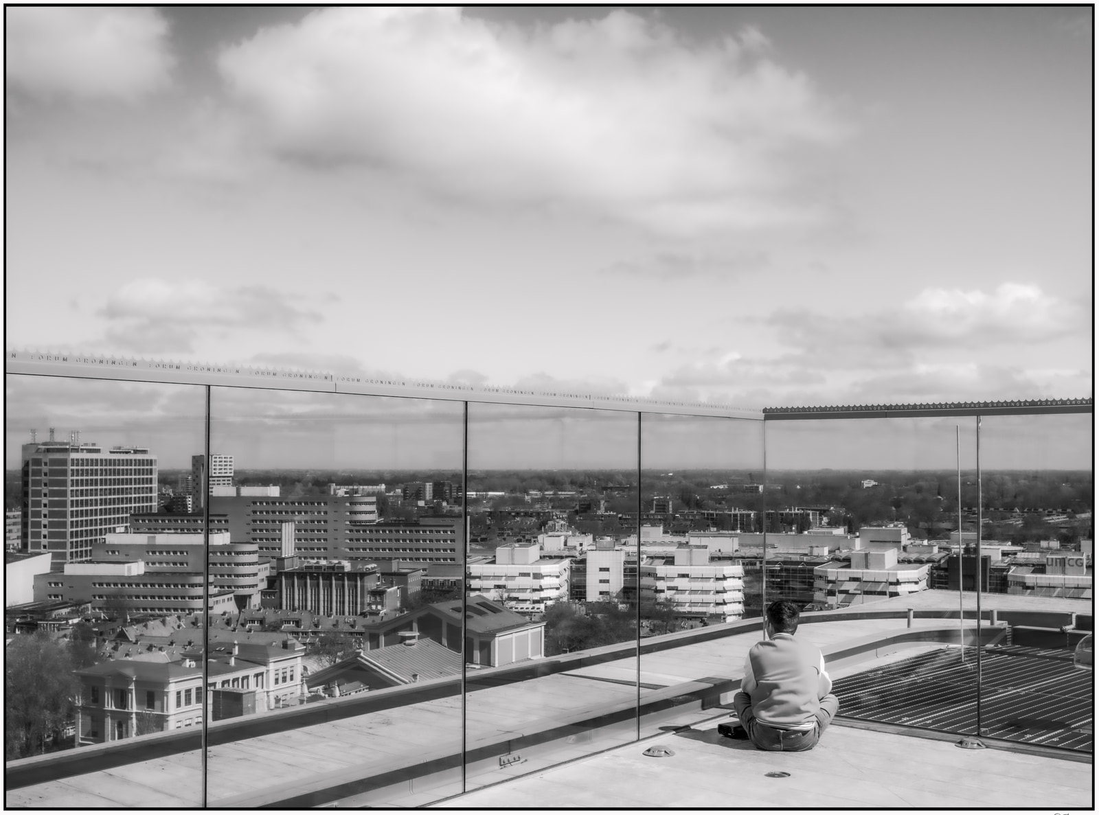 A person sits on a rooftop edge, gazing out over a cityscape framed by a large glass barrier.