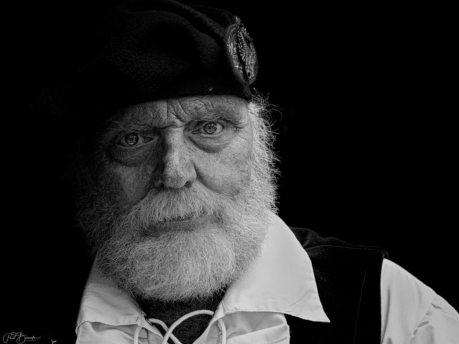 An elderly man with a long beard and mustache gazes directly at the camera, wearing a dark hat and a light-colored shirt.