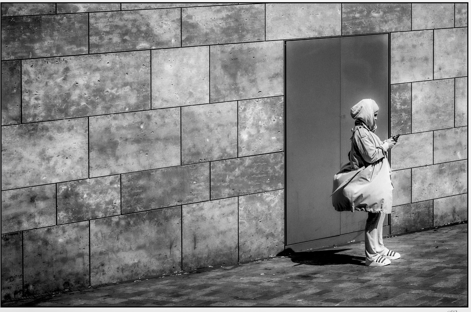 A person stands against a textured concrete wall, holding a phone, with a large bag slung over one shoulder.