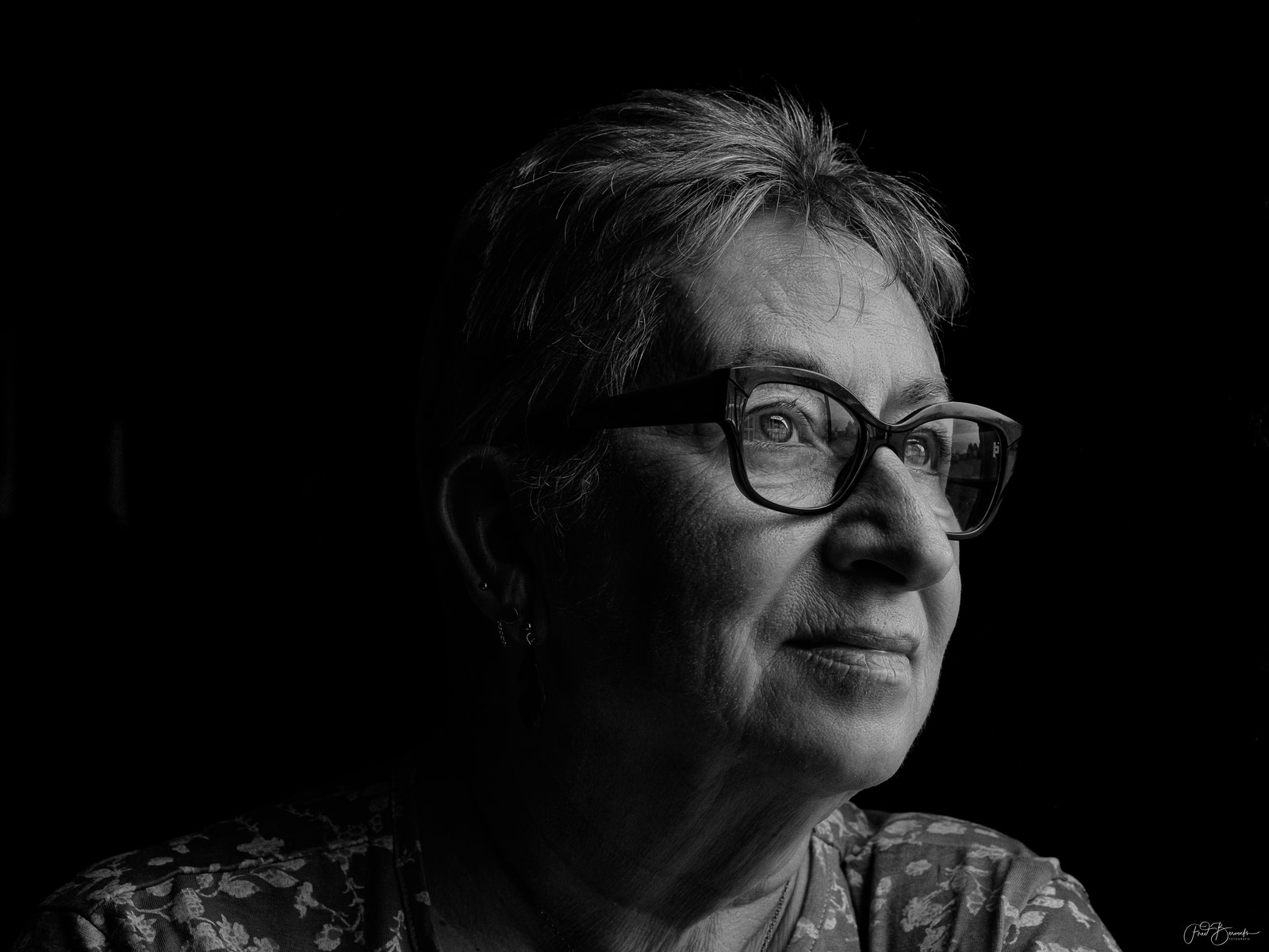 An older woman with glasses looks to the side against a dark background.