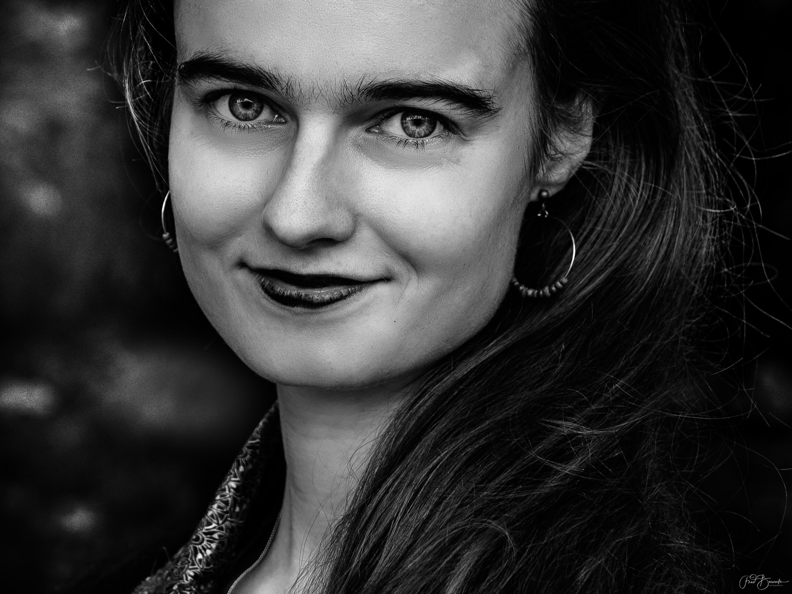 A close-up portrait of a person with long hair and hoop earrings, smiling gently against a dark background.