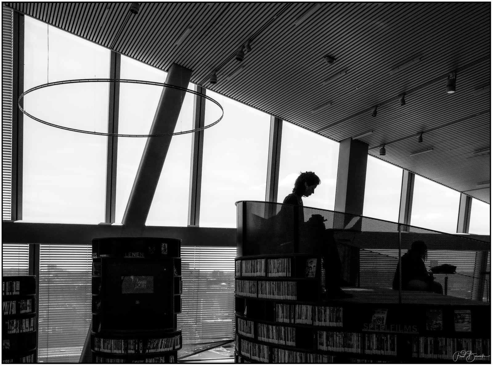 A person sits at a table in a modern interior with large windows, surrounded by bookshelves.