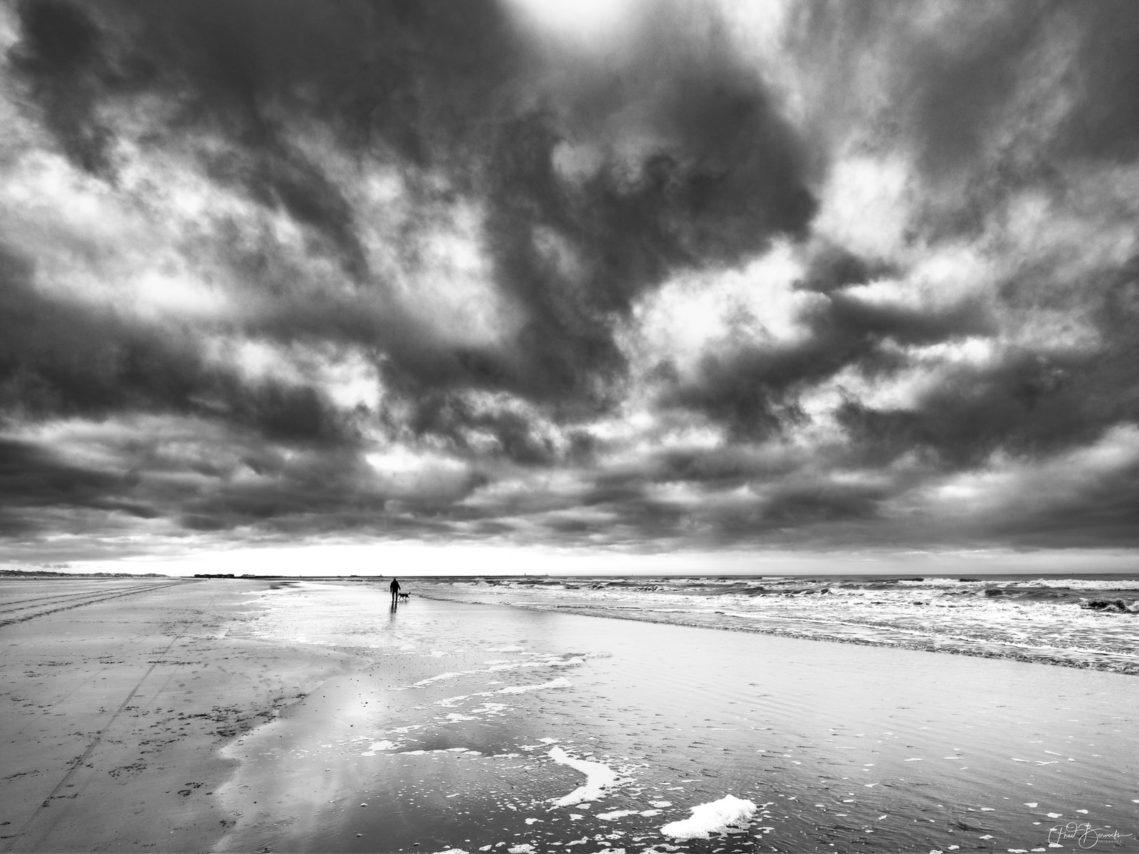 A lone figure walks a dog along a wide, empty beach under a dramatic, brooding sky.