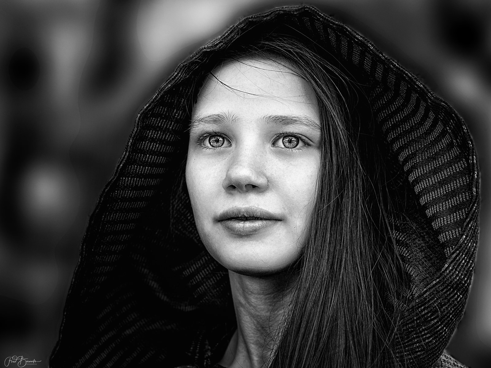 A person wearing a hooded garment gazes directly at the camera.