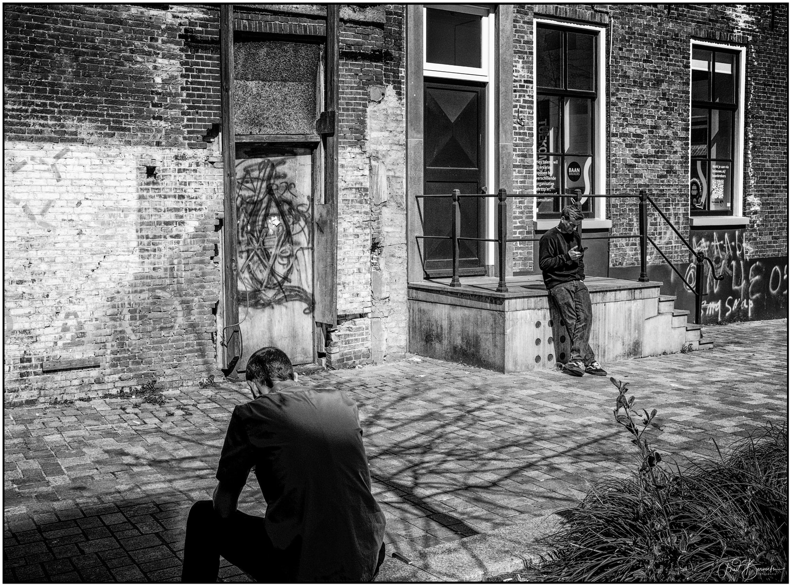 Two men sit on a brick-paved sidewalk in front of a weathered building with graffiti and windows; one leans against the building steps while the other crouches nearby.