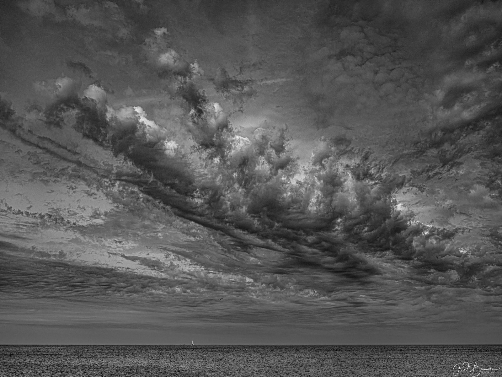 A dramatic black-and-white seascape shows turbulent, billowing clouds dominating the sky above a calm, expansive ocean.