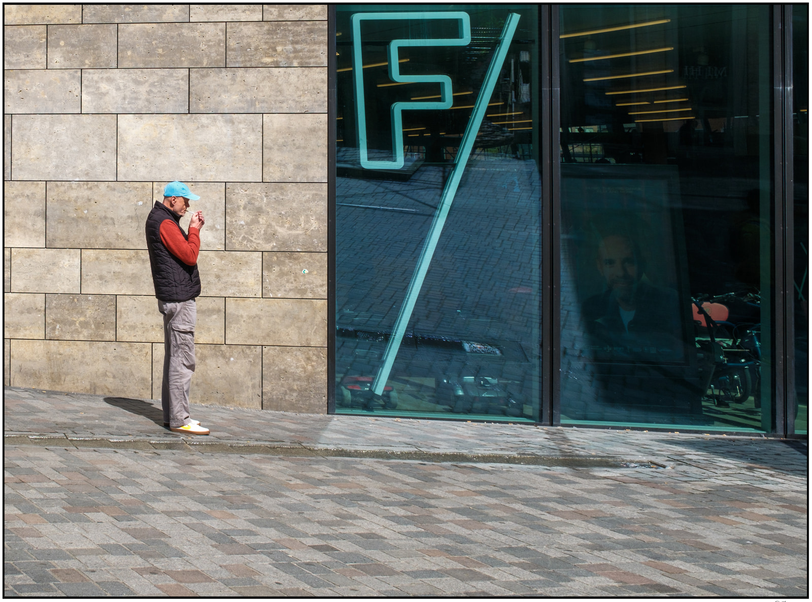 A man in a red shirt and black vest stands near a building entrance, looking at his phone.